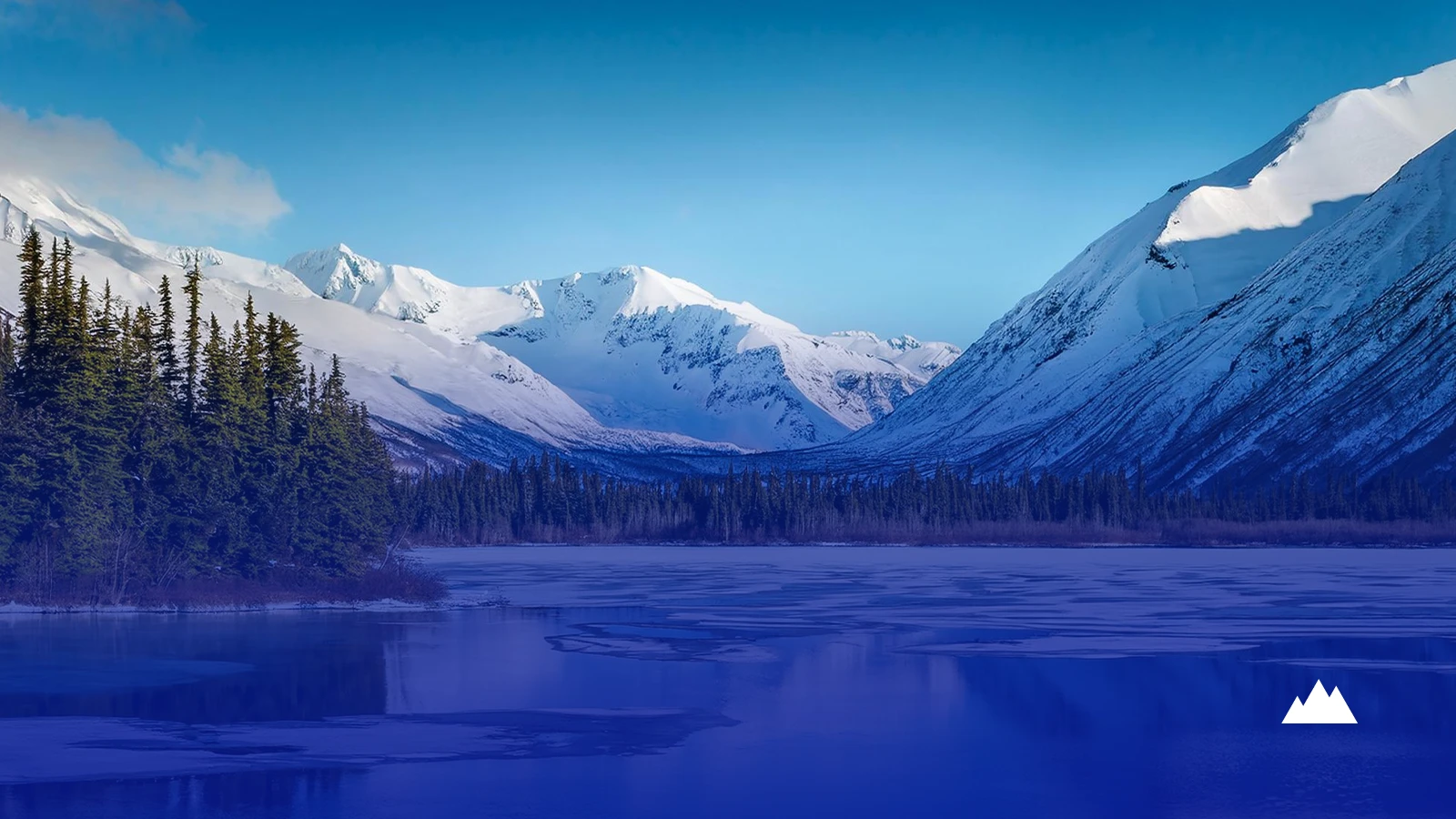 A frozen lake below snow-covered peaks, with spruce forest along the shoreline